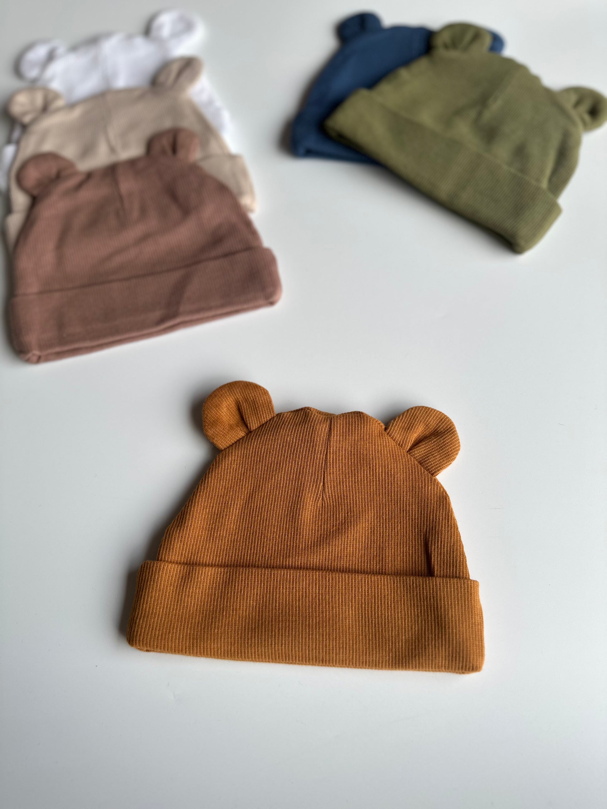 Infant bear hat in burnt orange, laid out on white background with other newborn hats in the background
