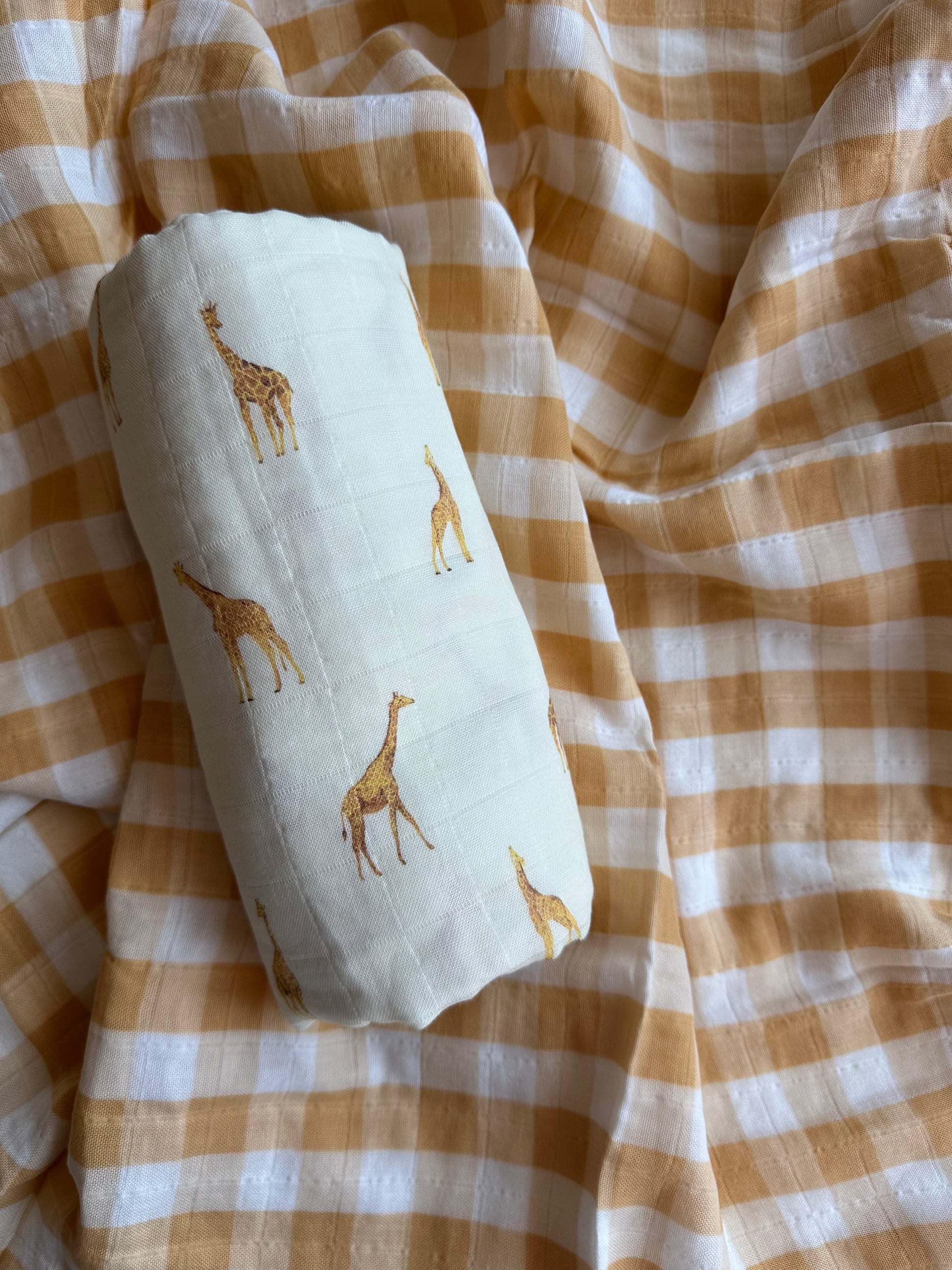 Giraffe muslin swaddle blanket rolled up and laid on top of yellow checkered blanket