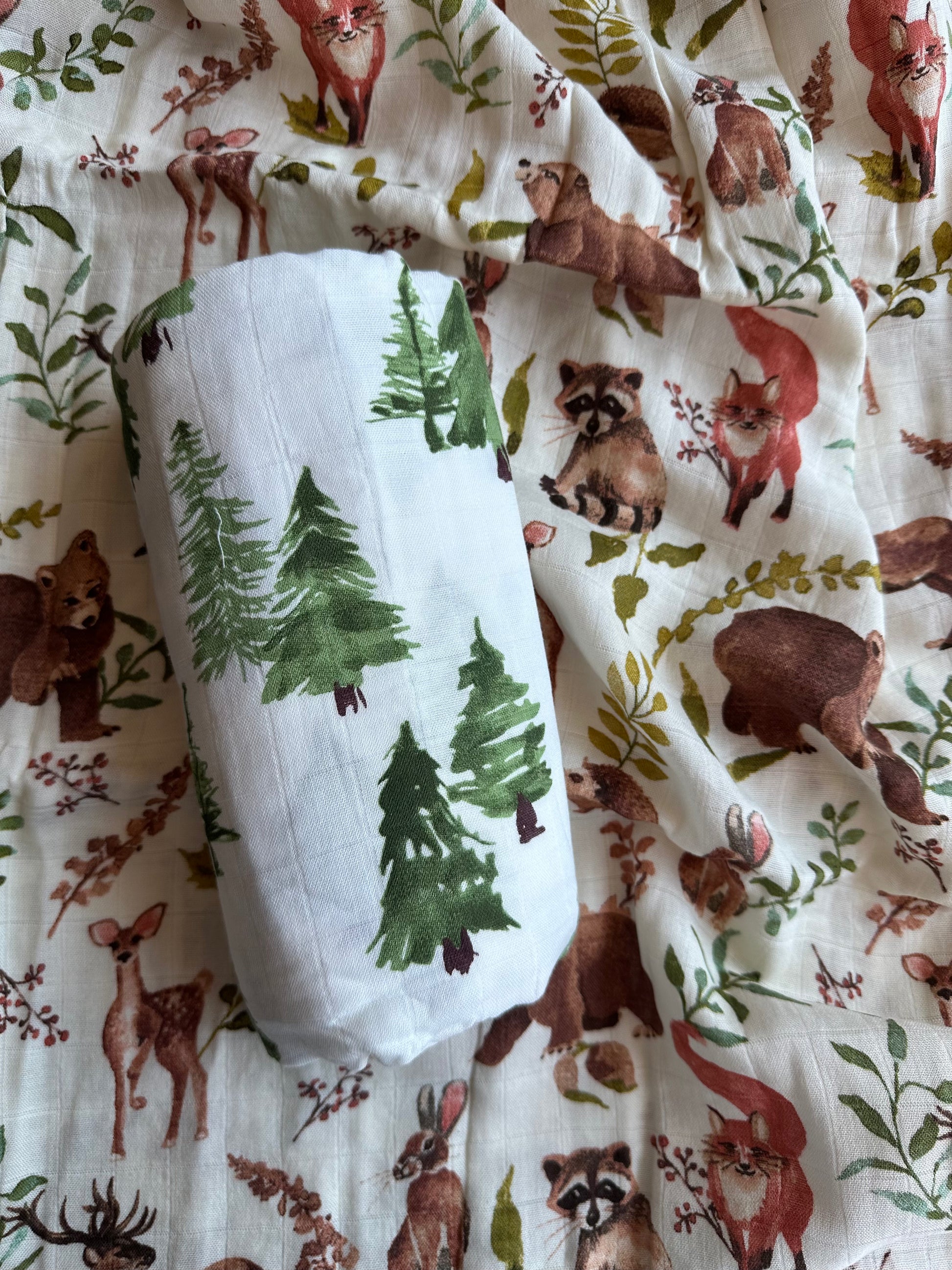 Tree muslin swaddle blanket rolled up and placed on top of woodland print muslin blanket