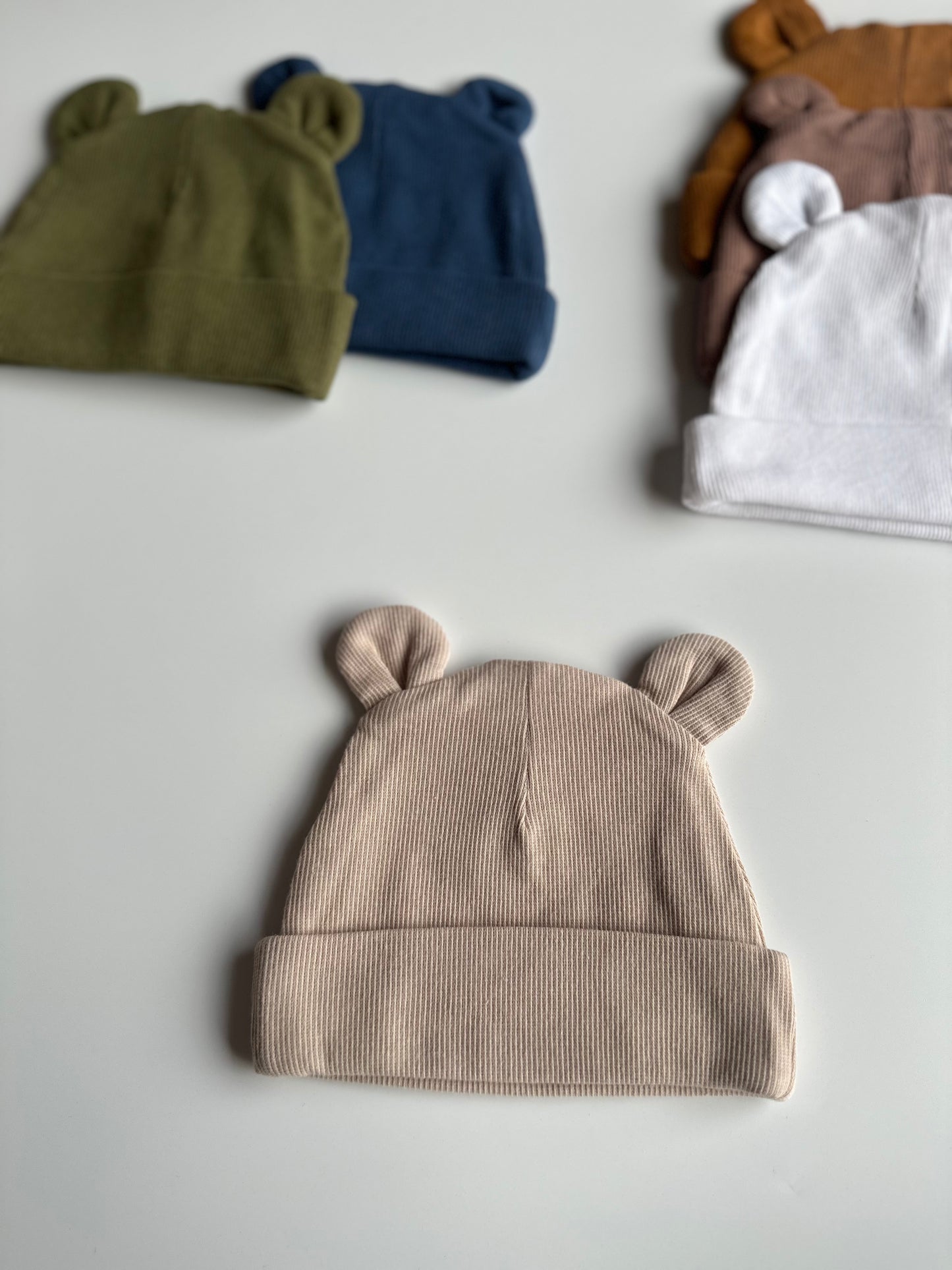 Infant bear hat in oat, laid out on white background with other newborn hats in the background