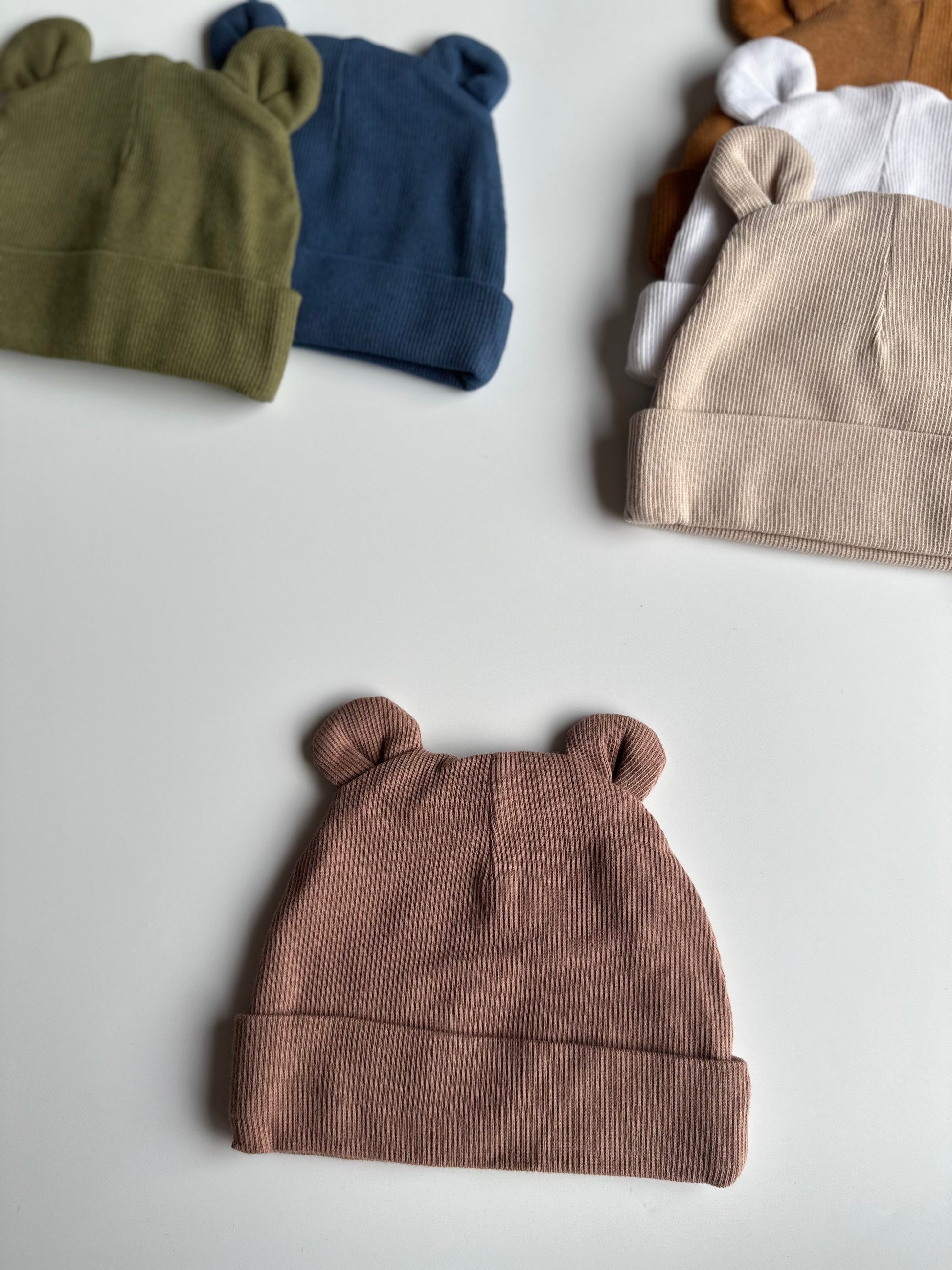 Infant bear hat in taupe, laid out on white background with other newborn hats in the background