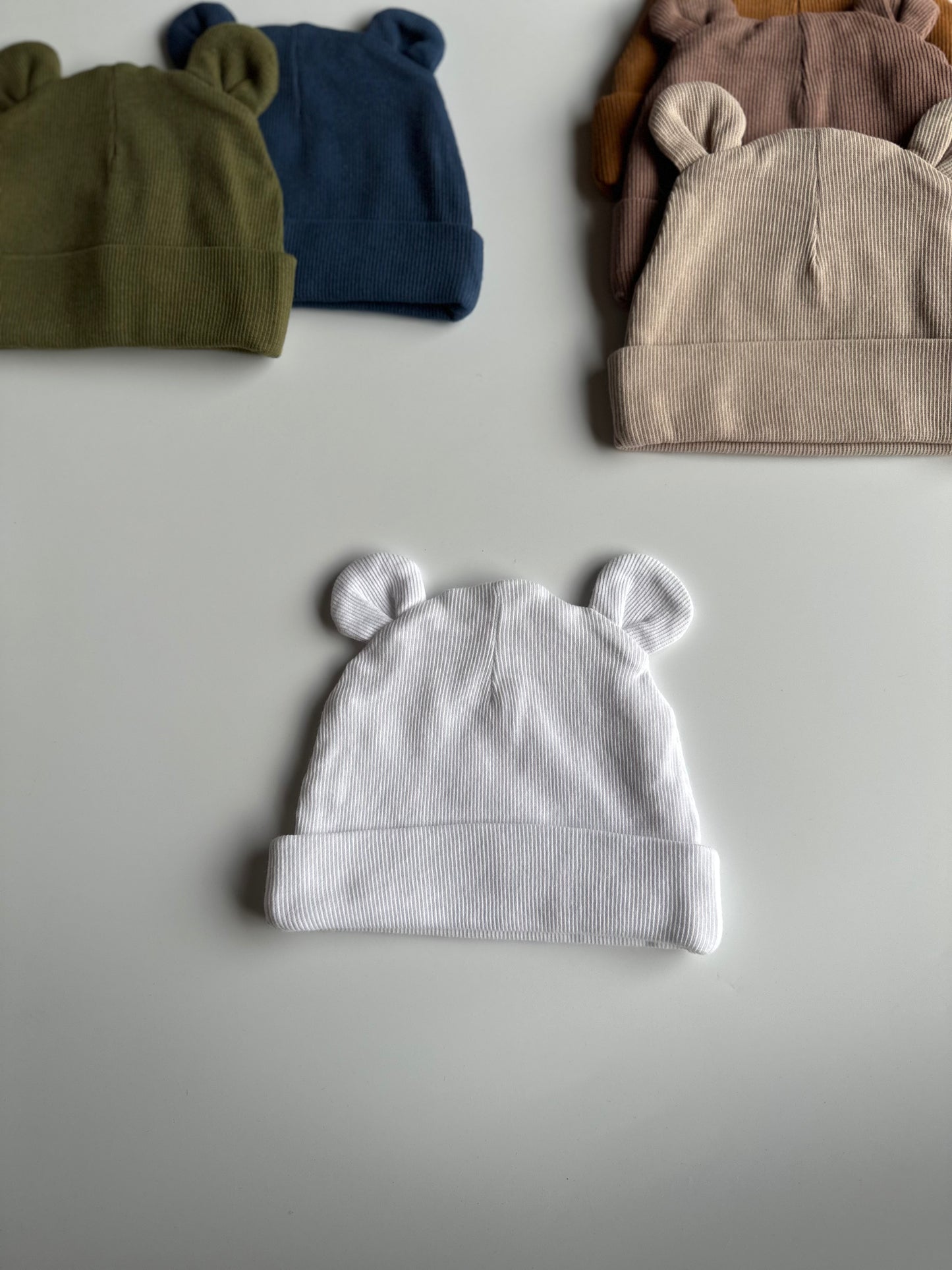 Infant bear hat in white, laid out on white background with other newborn hats in the background