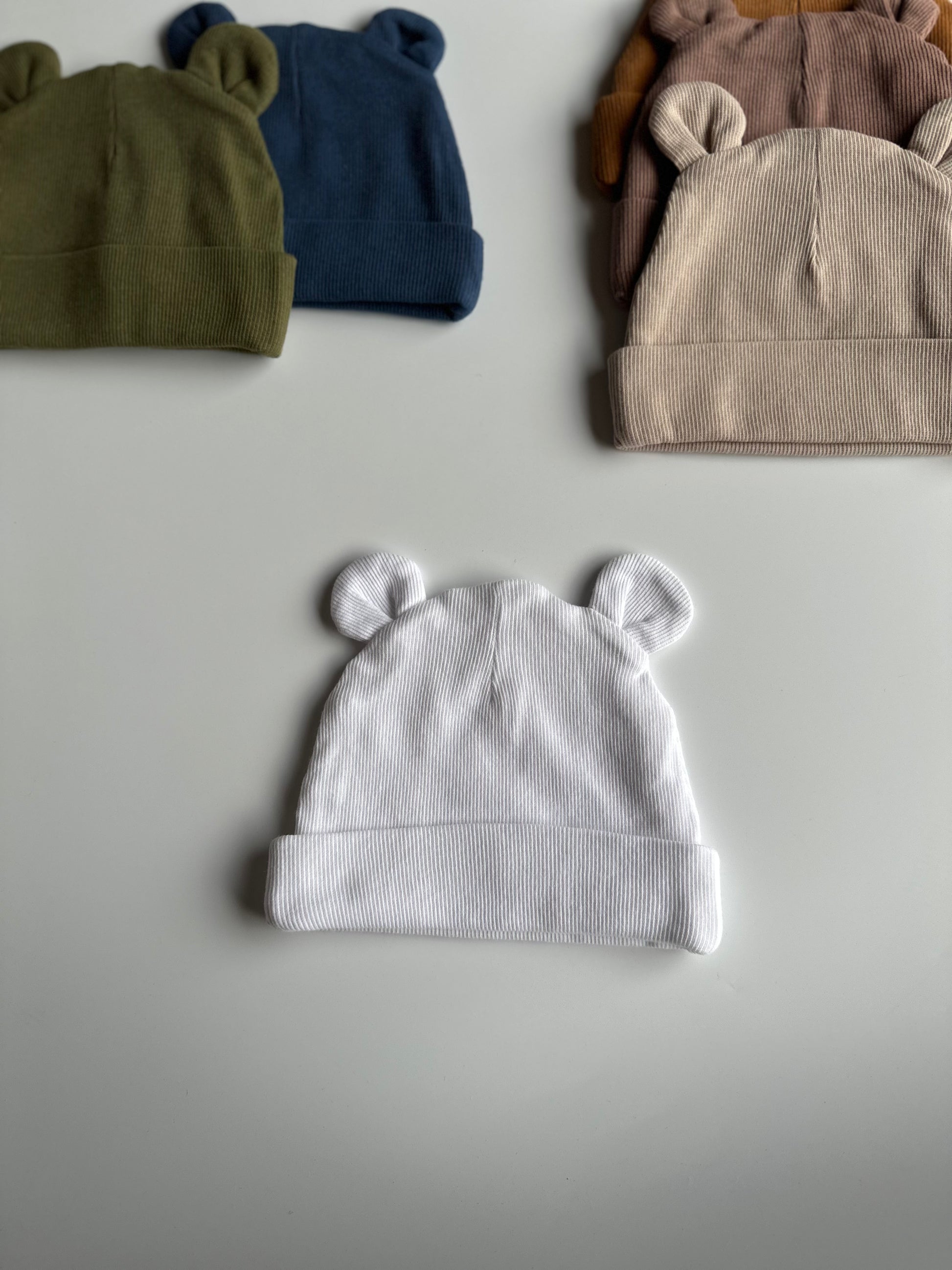 Infant bear hat in white, laid out on white background with other newborn hats in the background